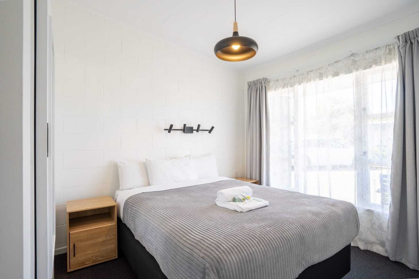 Ein modernes Hotelzimmer mit einem großen Bett, einem Nachttisch aus Holz, einer Pendelleuchte und einem Fenster mit Vorhängen.