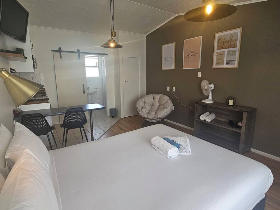 Une chambre d’hôtel avec un grand lit, un bureau avec une chaise, une chaise supplémentaire, une petite table avec un ventilateur et un mur orné de tableaux.