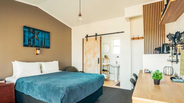 A modern hotel room with a bed, a desk, a sink, and a painting on the wall. - Frimley Lodge Motel (Hastings)