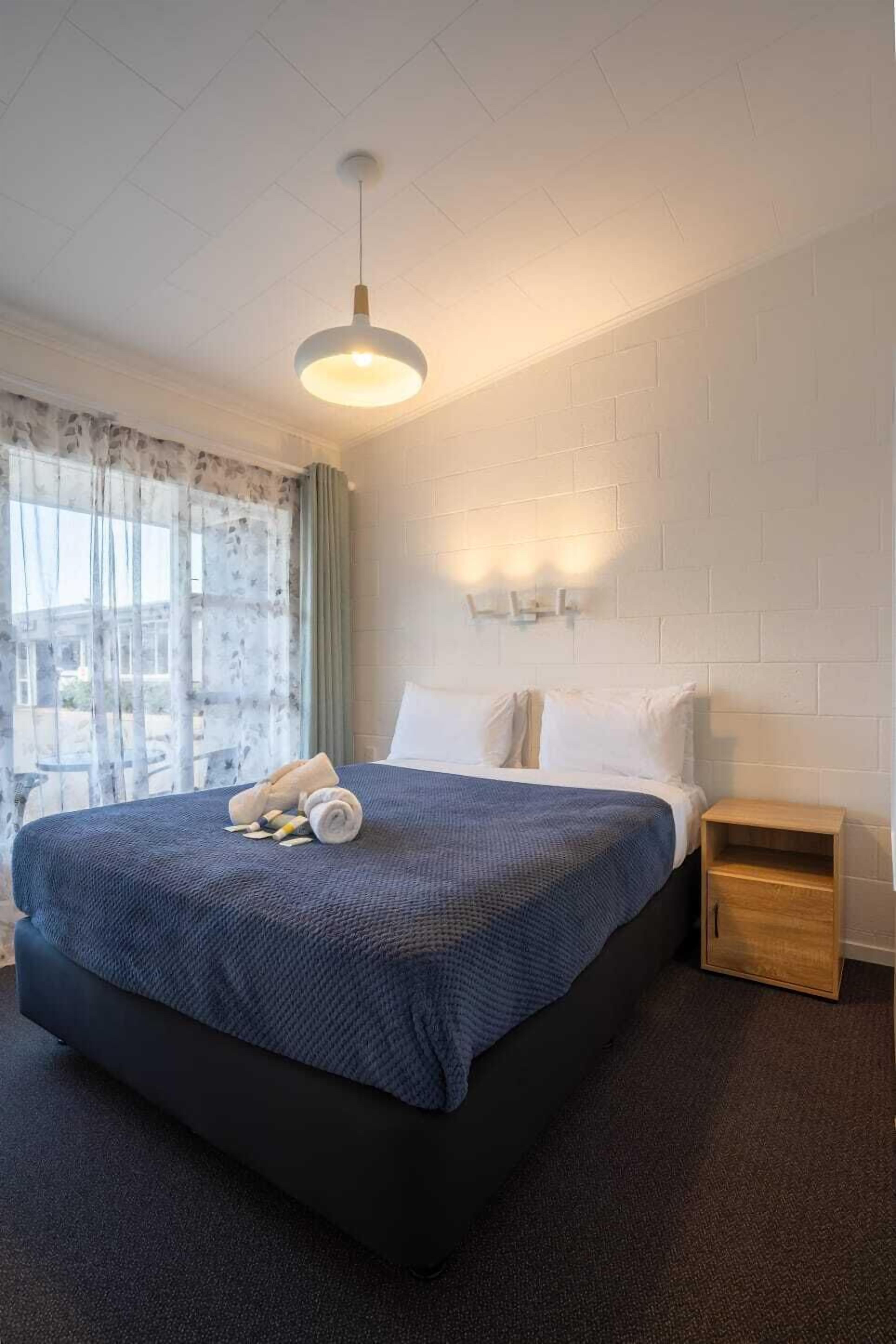 A hotel room with a large bed, a small wooden nightstand, a wall-mounted light fixture, and a window with sheer curtains.