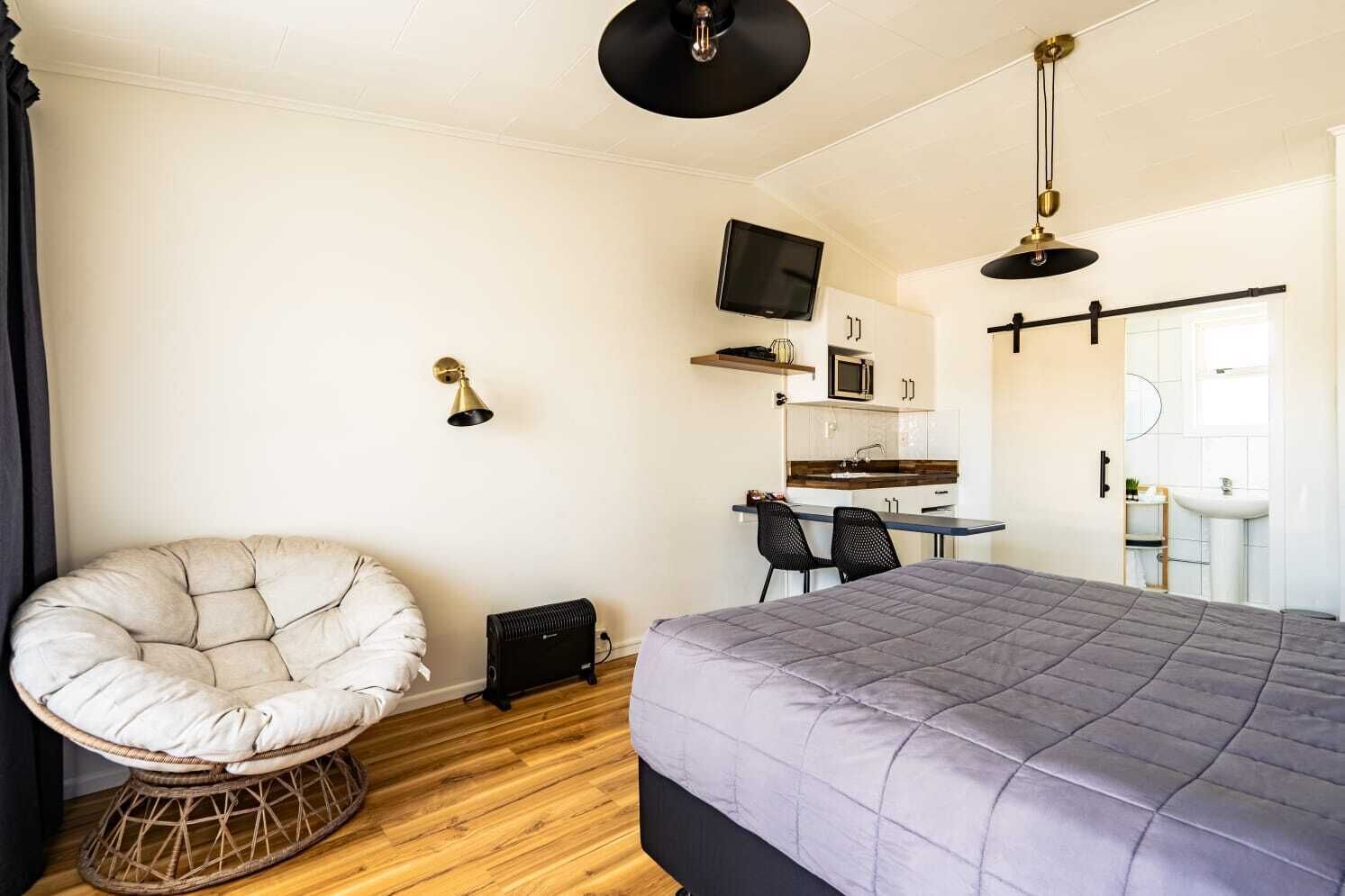 A compact room with a bed, a chair, a desk, a TV, and a kitchenette.