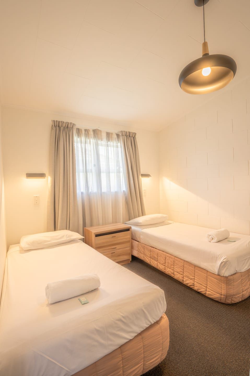 Habitación de hotel con dos camas, una mesita de noche de madera, una lámpara de pared y un ventanal con cortinas.