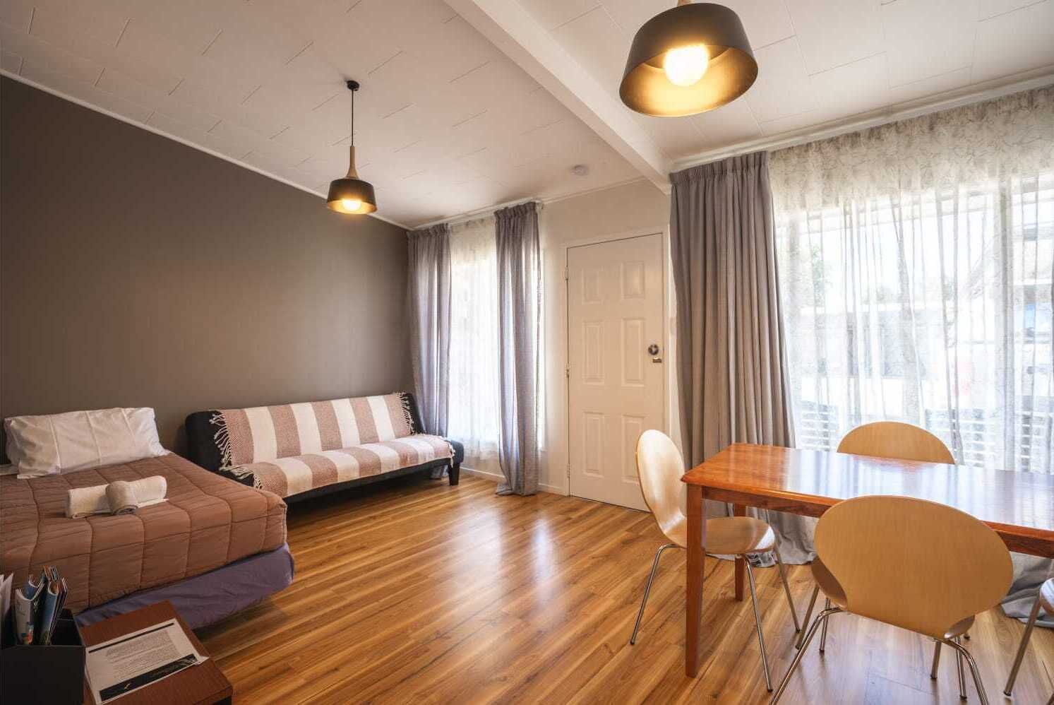 A modern living room with a sofa, a dining table with chairs, wooden flooring, and a large window with curtains.
