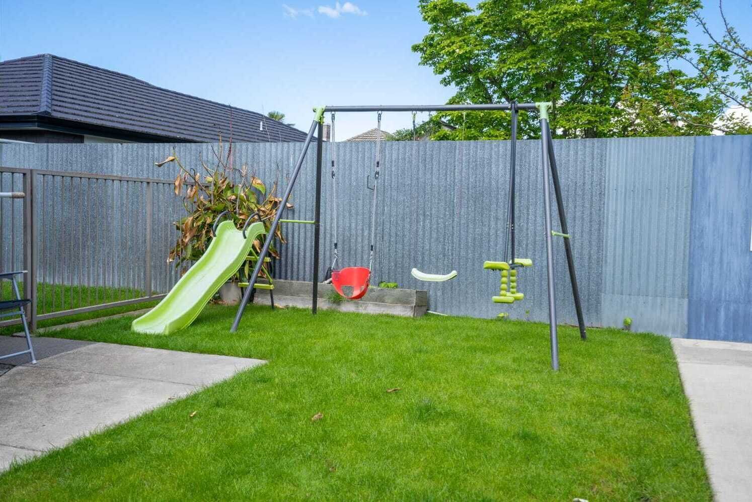 A backyard with a green slide, red swing, and yellow exercise equipment.