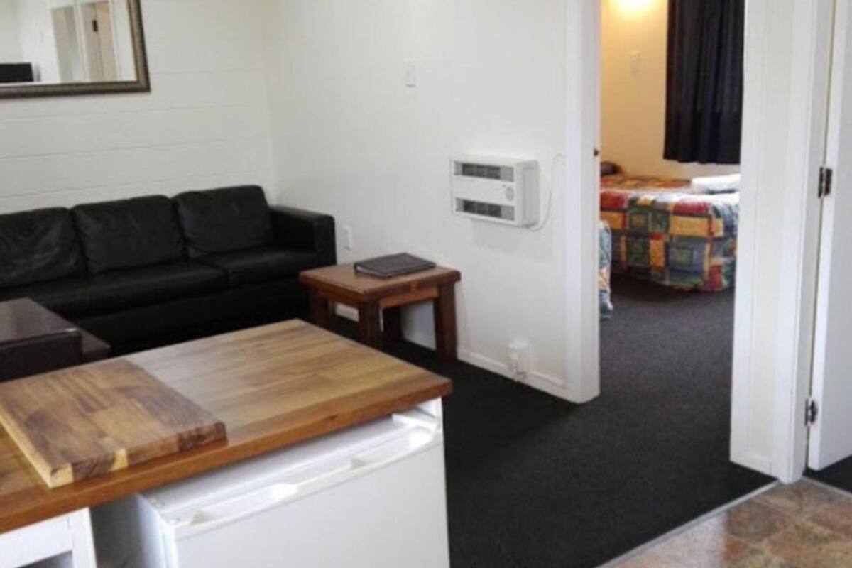 Een knus woongedeelte met een kitchenette, een zwarte bank, een kleine houten tafel en een doorgang naar een slaapkamer met een kleurrijke quilt.