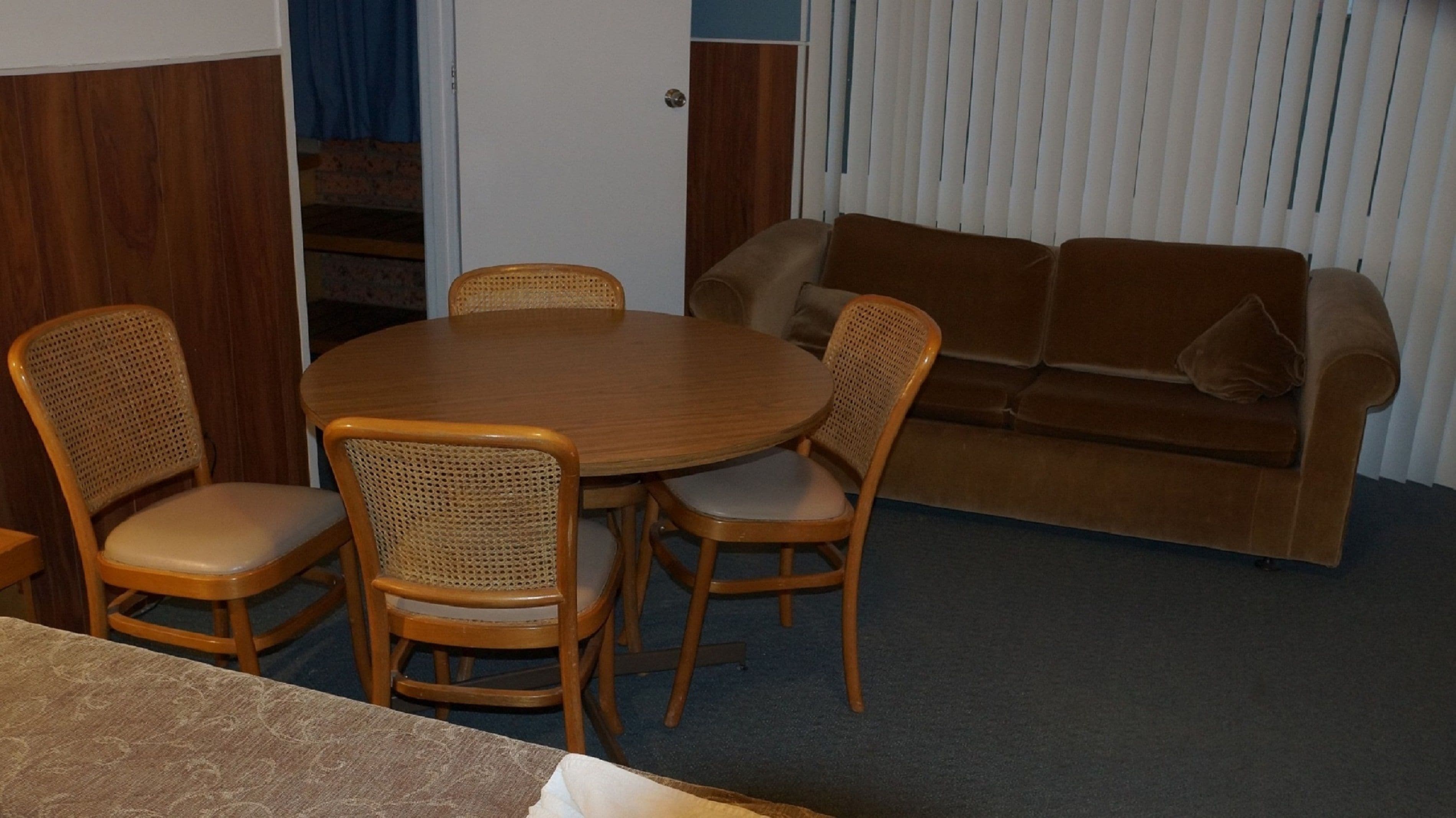 A room with a round wooden table surrounded by four wicker chairs, a brown sofa, and a door leading to a staircase.