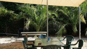 Un área de estar al aire libre con mesa y sillas bajo una estructura cubierta, rodeada de exuberante vegetación y palmeras.