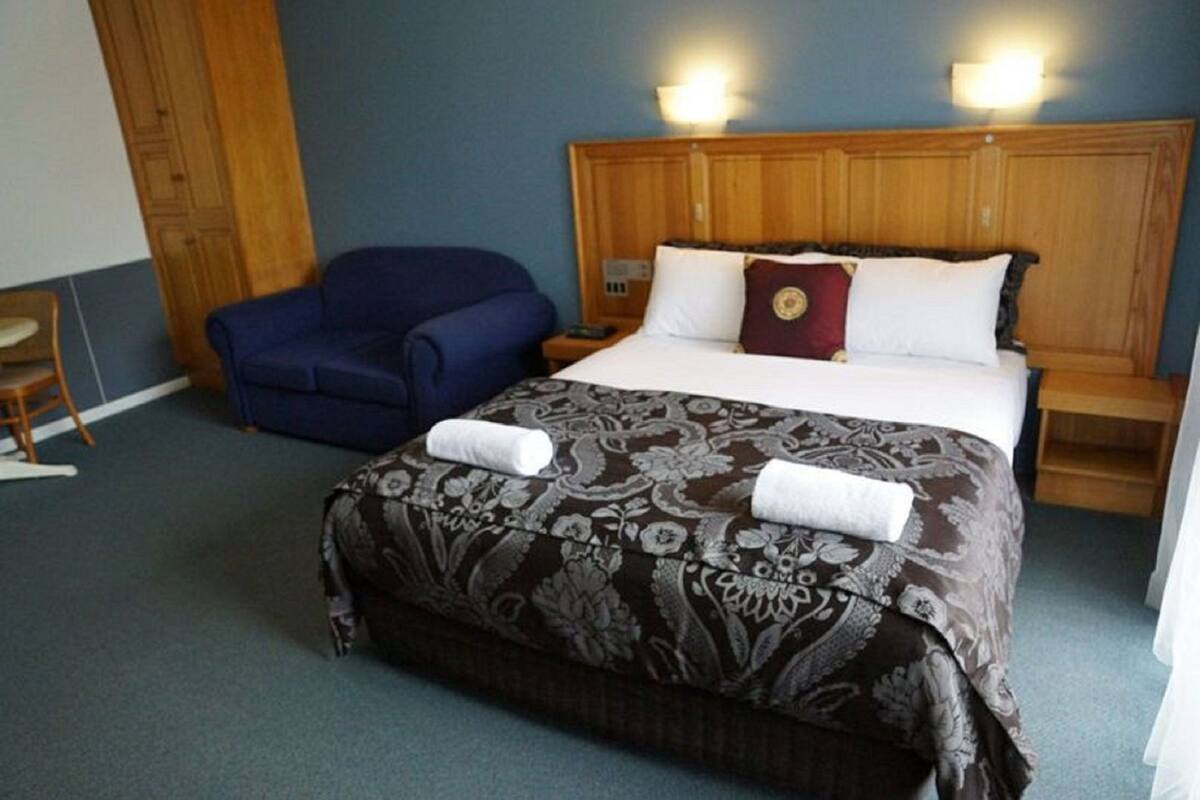 A hotel room with a large bed, a blue armchair, a wooden headboard, and a nightstand with a lamp.