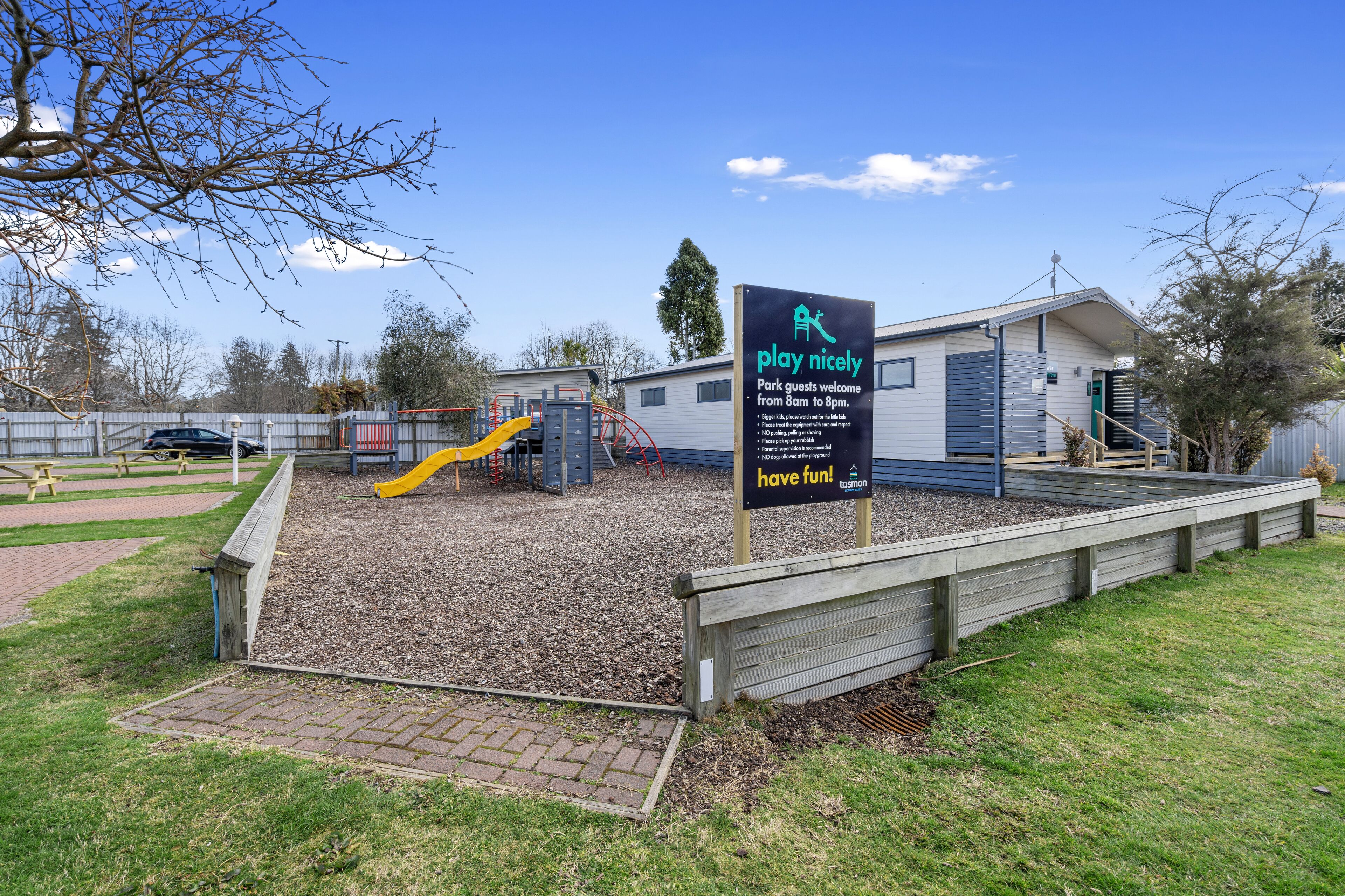 A playground area with a slide and play structures, a picnic table, and a sign welcoming park guests.