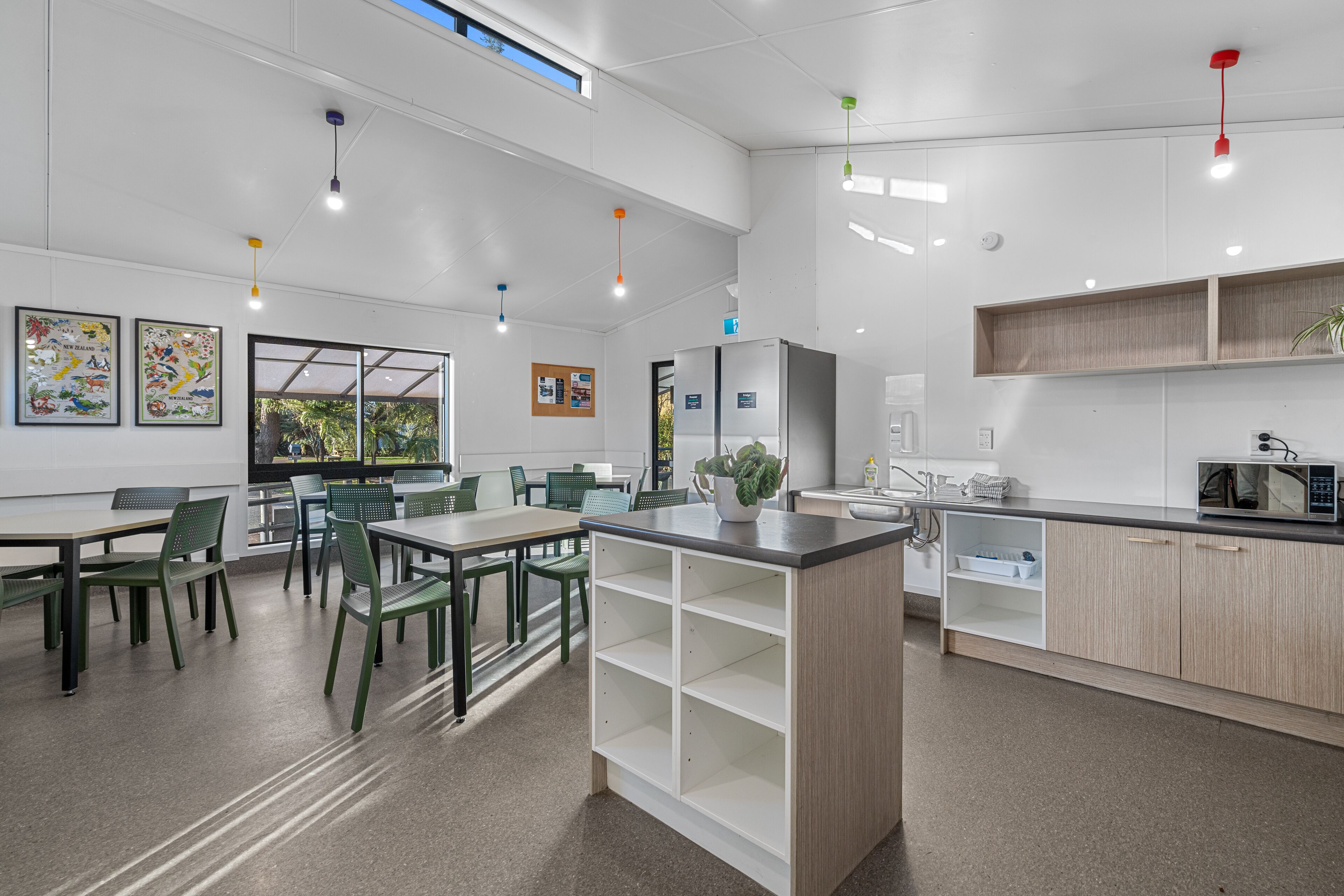 A modern kitchen with white cabinets, a central island, and a microwave. There are green chairs around a table, and a window with a view of greenery.