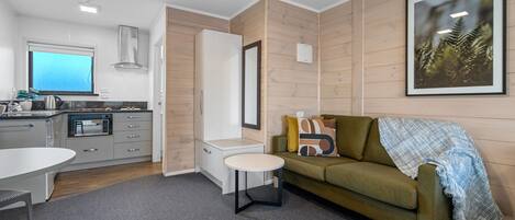 A modern living room with a green sofa, a small round table, and a kitchen area with a microwave and kettle.