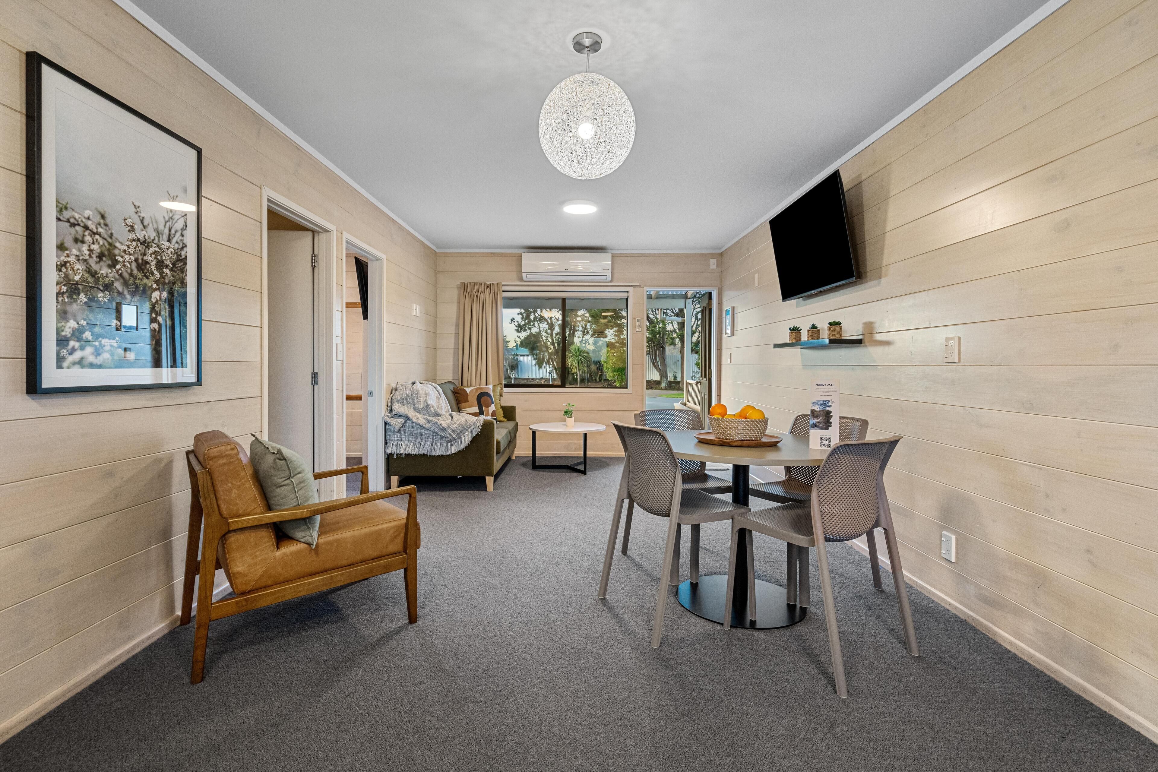 A modern living room with a sofa, a dining table with chairs, a TV mounted on the wall, and a ceiling light fixture.