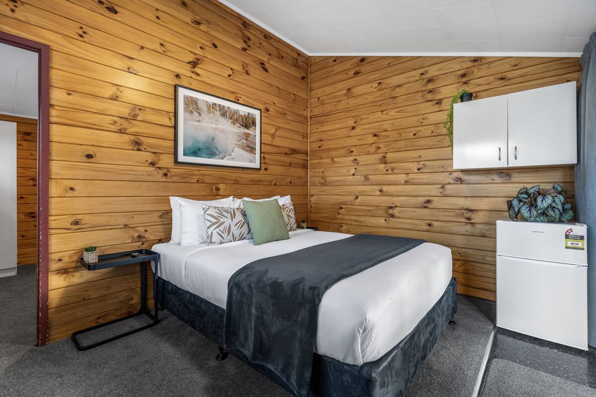 Una habitación con un revestimiento de madera, una cama con ropa de cama blanca y varias almohadas, un pequeño armario blanco y una fotografía de un paisaje nevado.