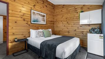 A room with a wooden paneled wall, a bed with white bedding and multiple pillows, a small white cabinet, and a picture of a snowy landscape.