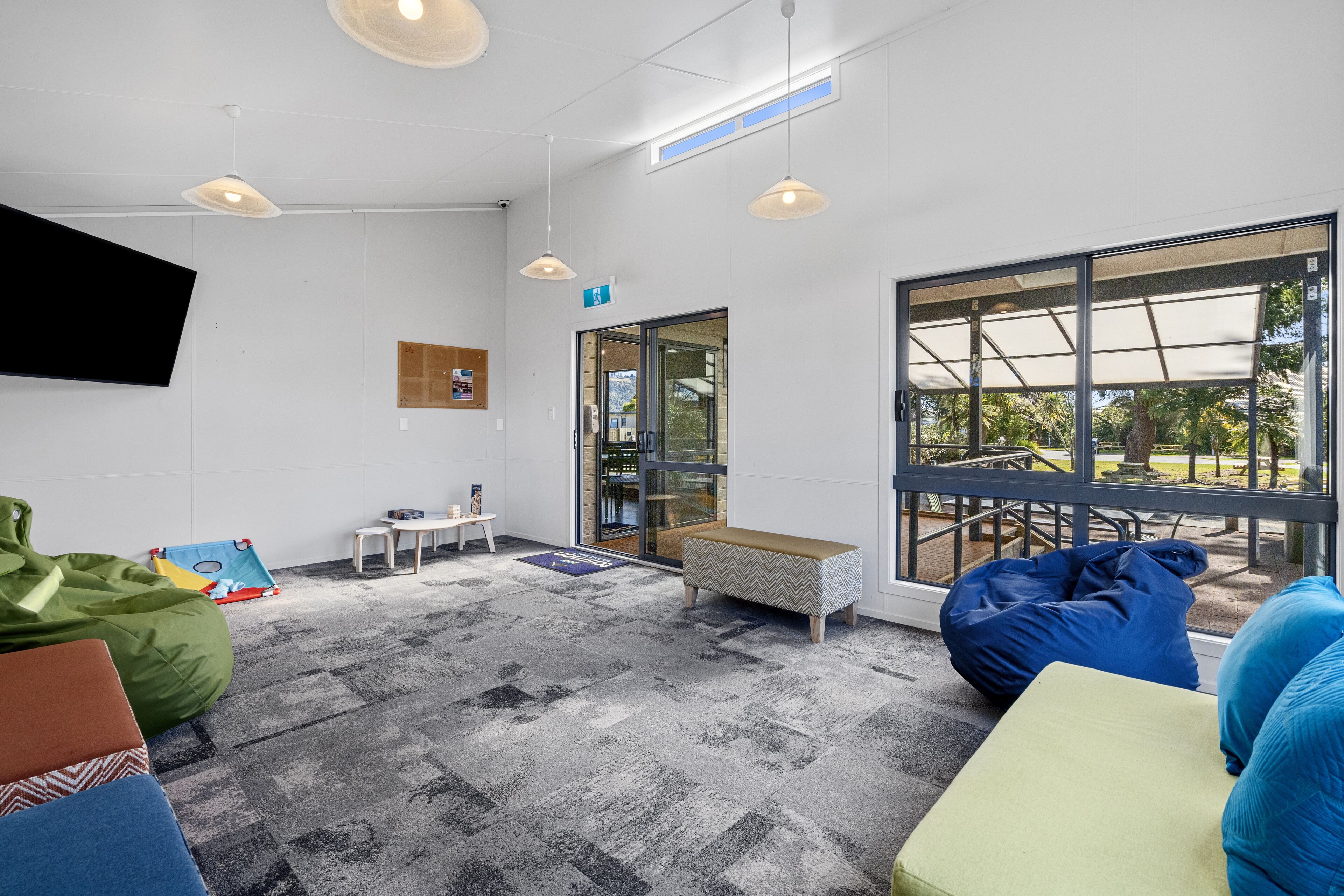 A modern, well-lit room with a large window, a flat-screen TV mounted on the wall, and a variety of seating options including bean bags and a bench.