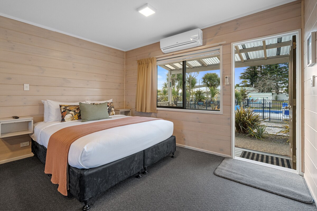A bedroom with a bed, a bedside table, a window, and a door leading to an outdoor area.