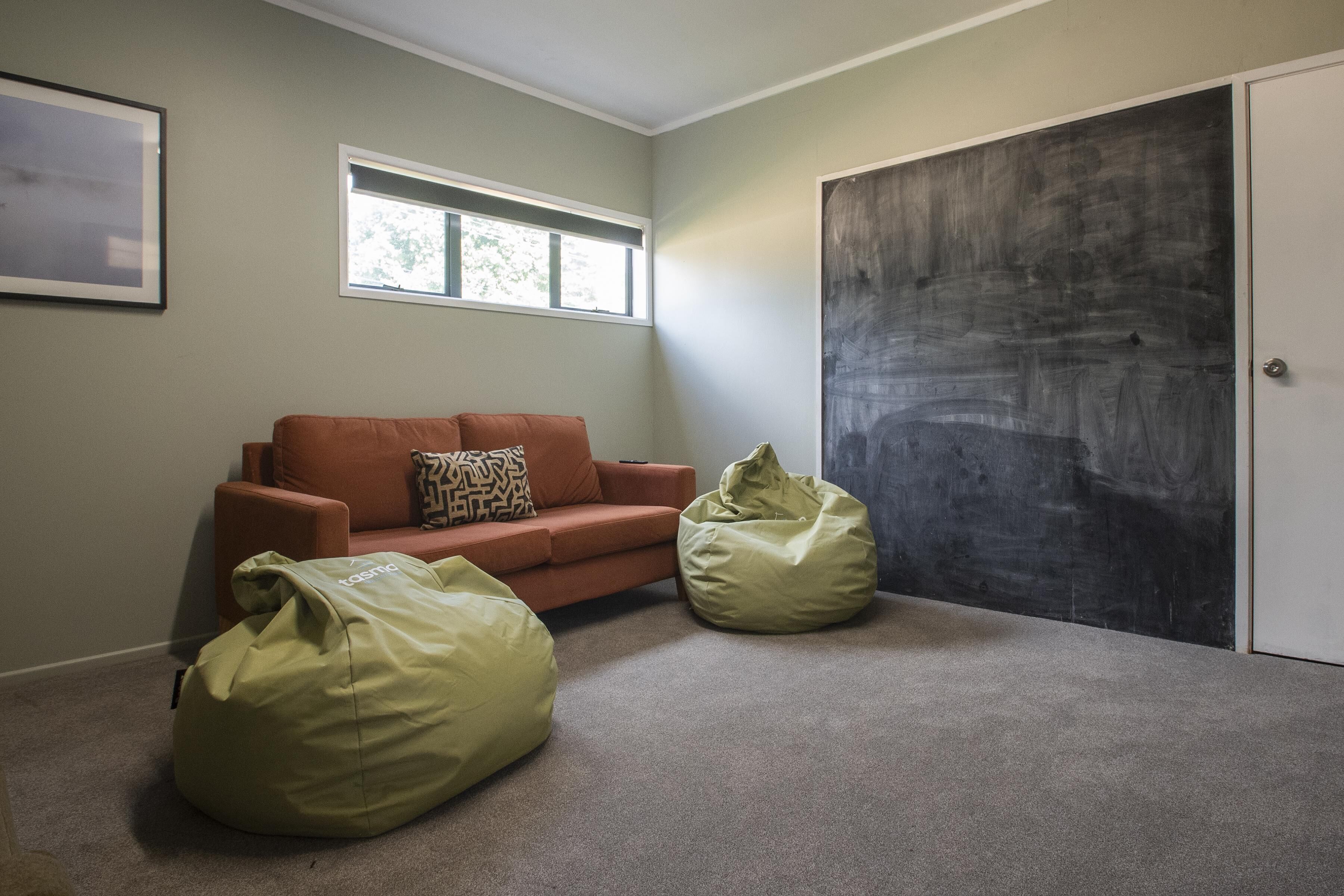 A room with a beige carpet, a brown sofa with patterned cushions, two green bean bags, a window, and a door with a dark textured panel.