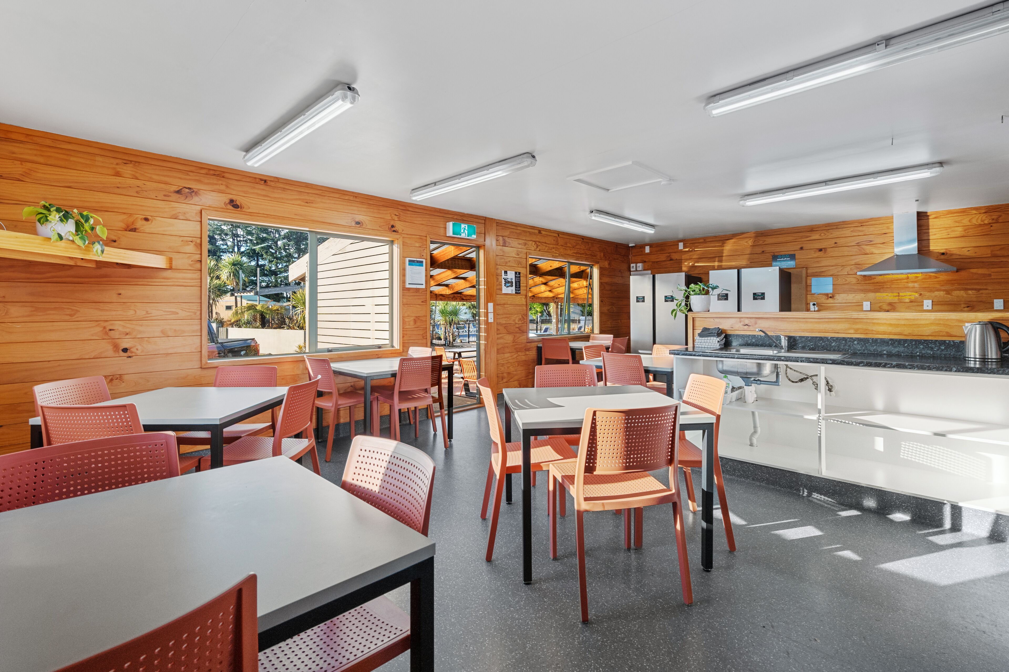 A modern restaurant with wooden walls, multiple tables and chairs, a kitchen area, and a view of the outdoors.