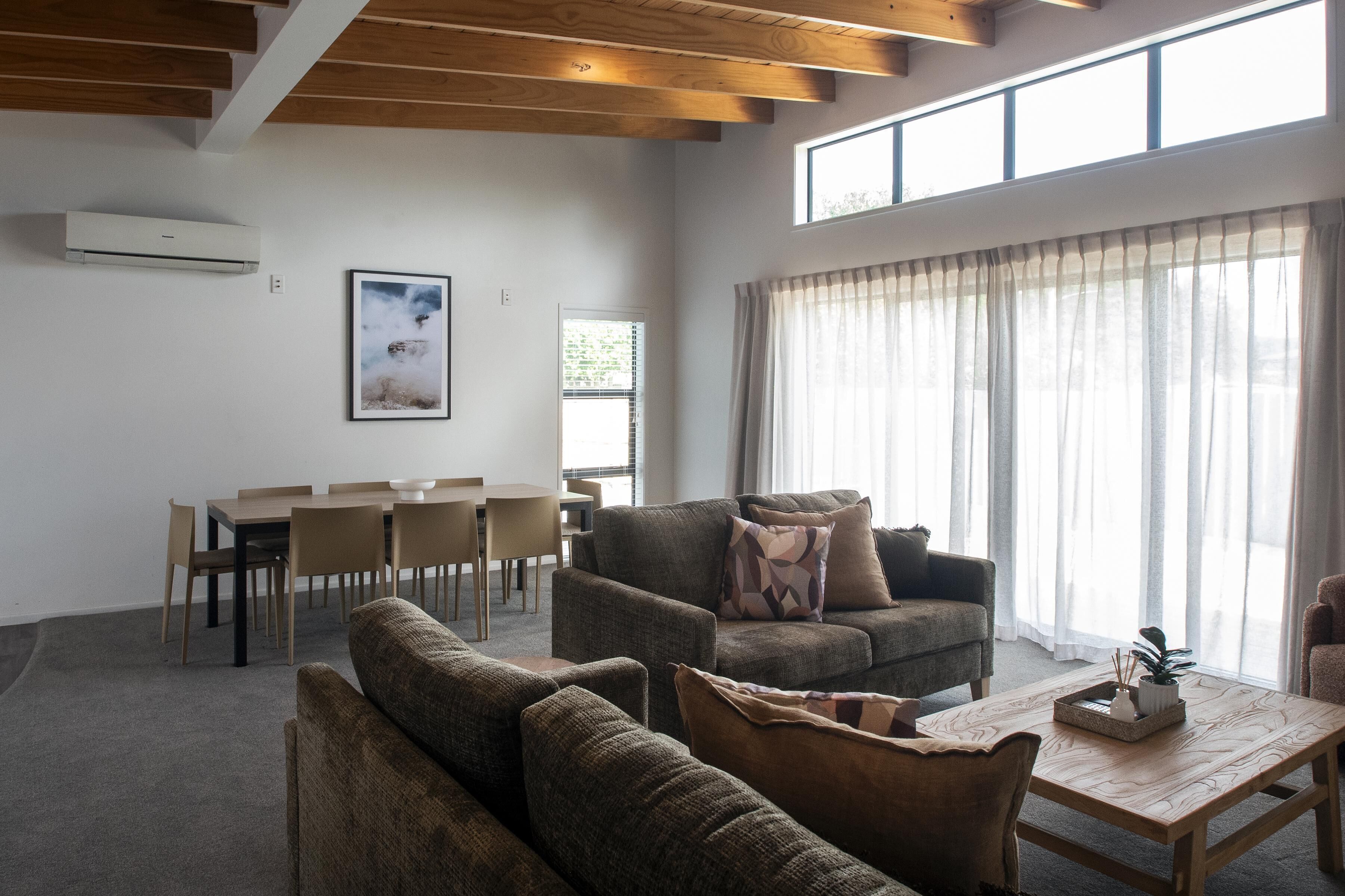 A spacious living room with a wooden ceiling, a dining area with chairs and a table, and large windows with sheer curtains.