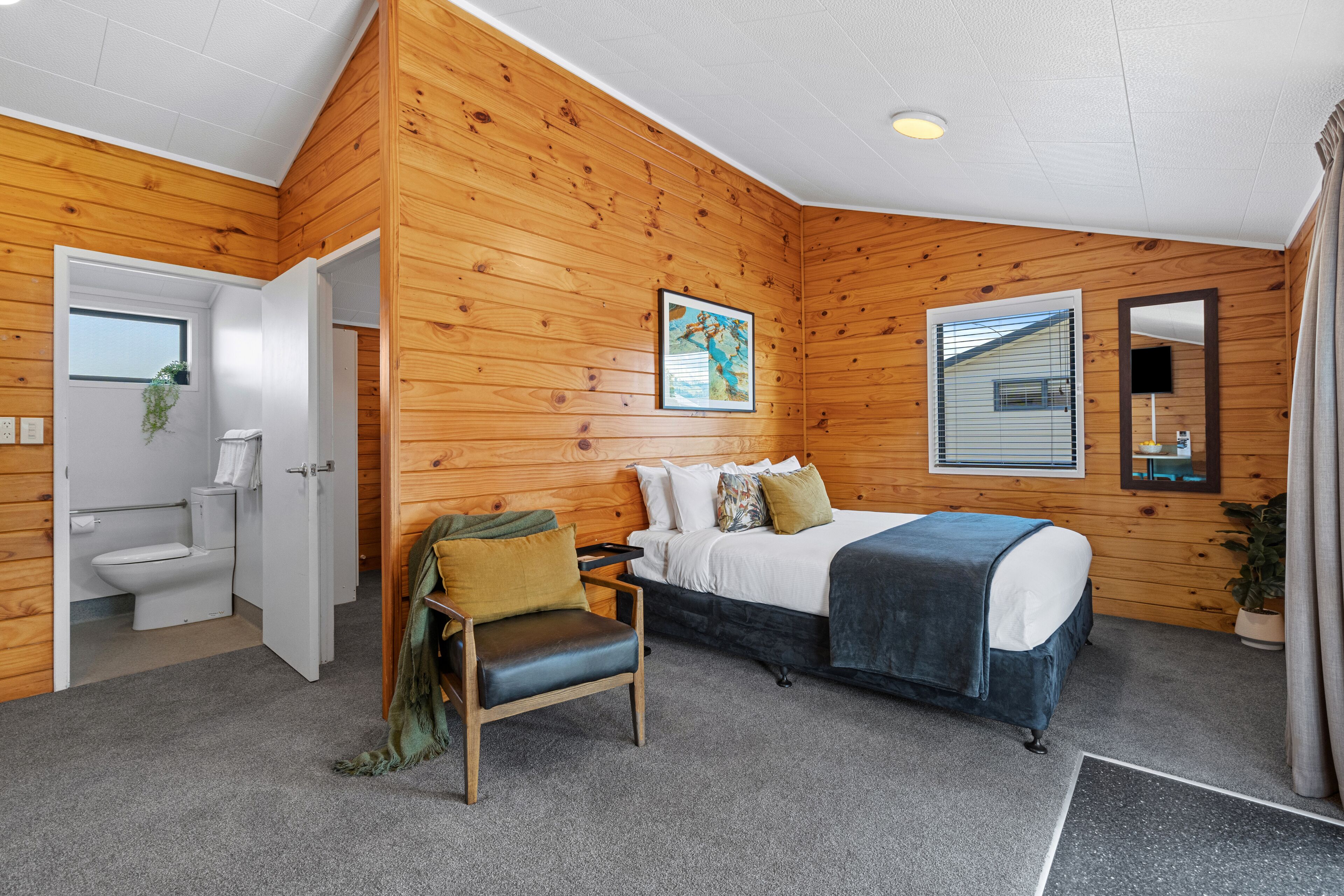 A room with wooden walls, a bed with a blue blanket, a chair, a bedside table, a window with blinds, a mirror, and a plant.