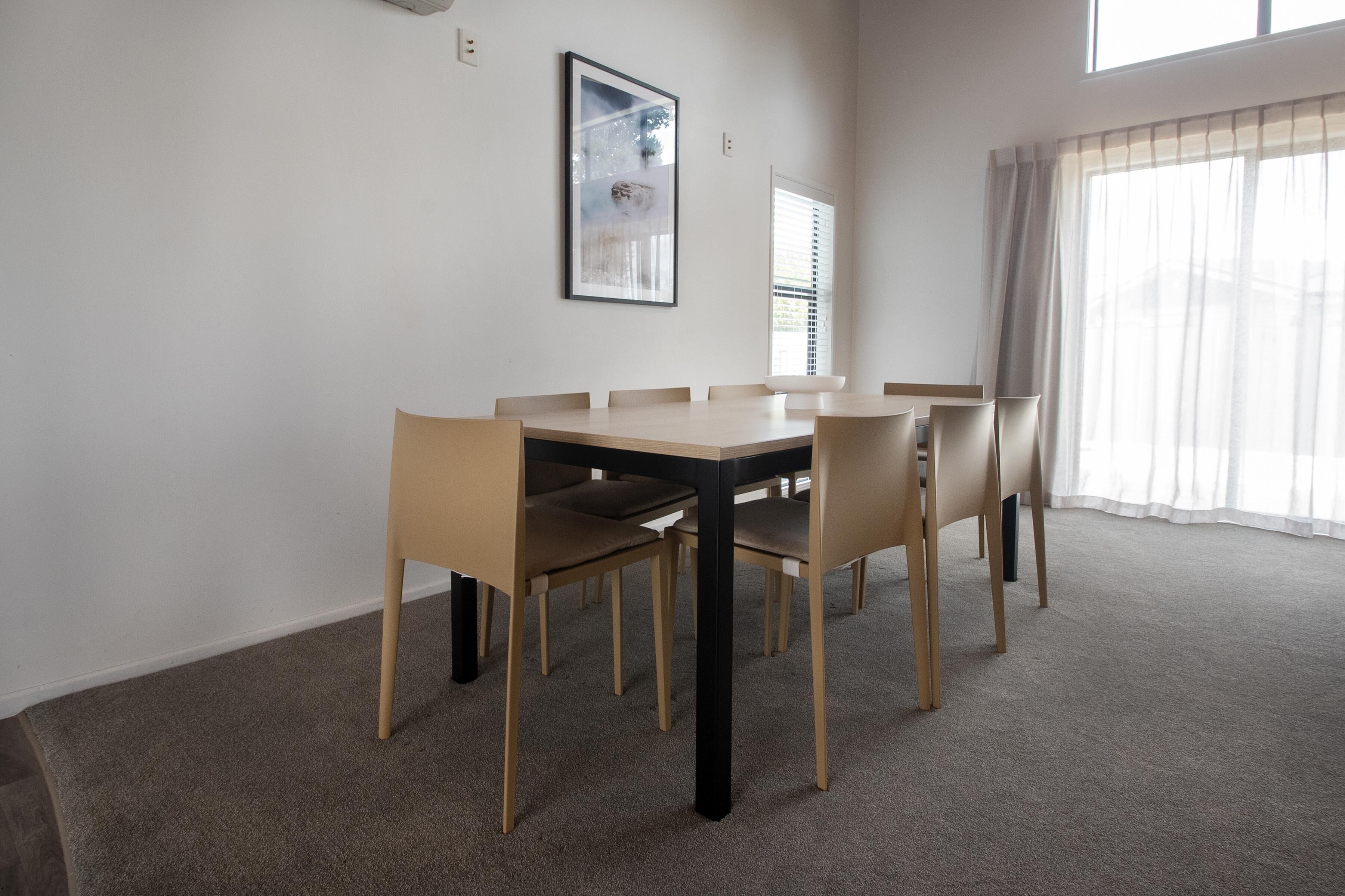 A dining area with a rectangular table and six chairs, a framed picture on the wall, and a window with blinds.