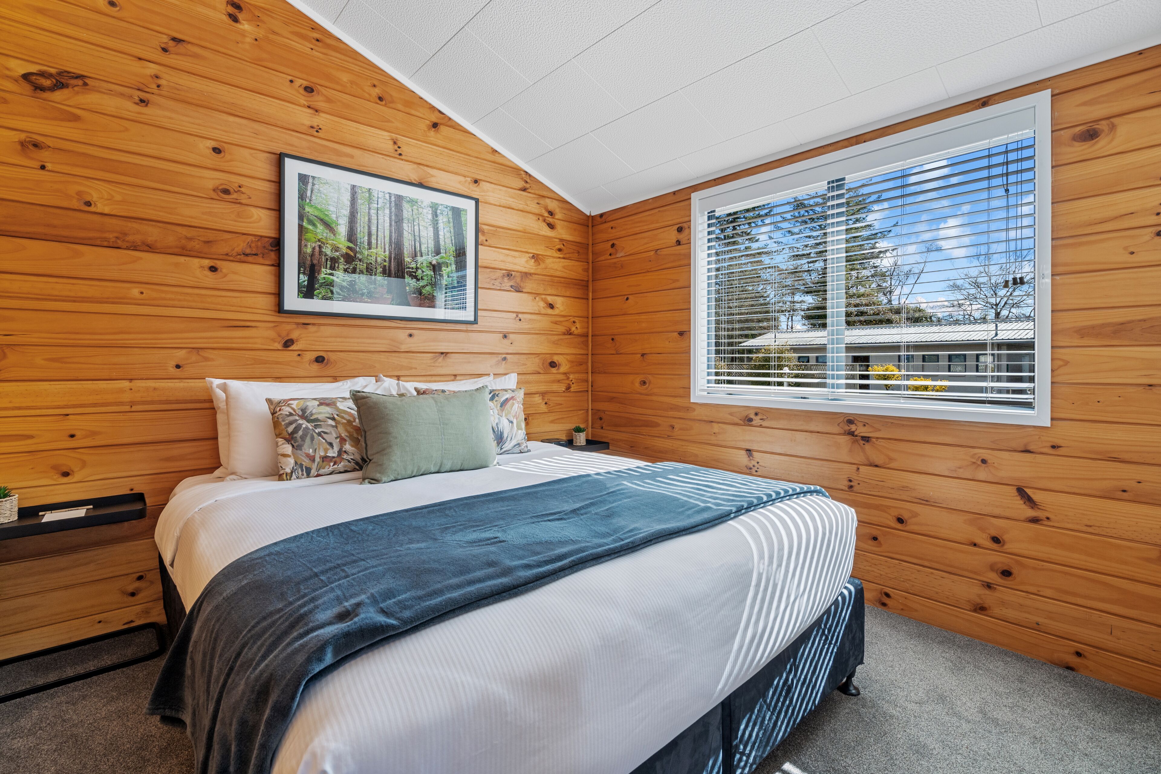 A bedroom with wooden walls, a bed with a blue blanket, a framed picture on the wall, and a window with blinds.