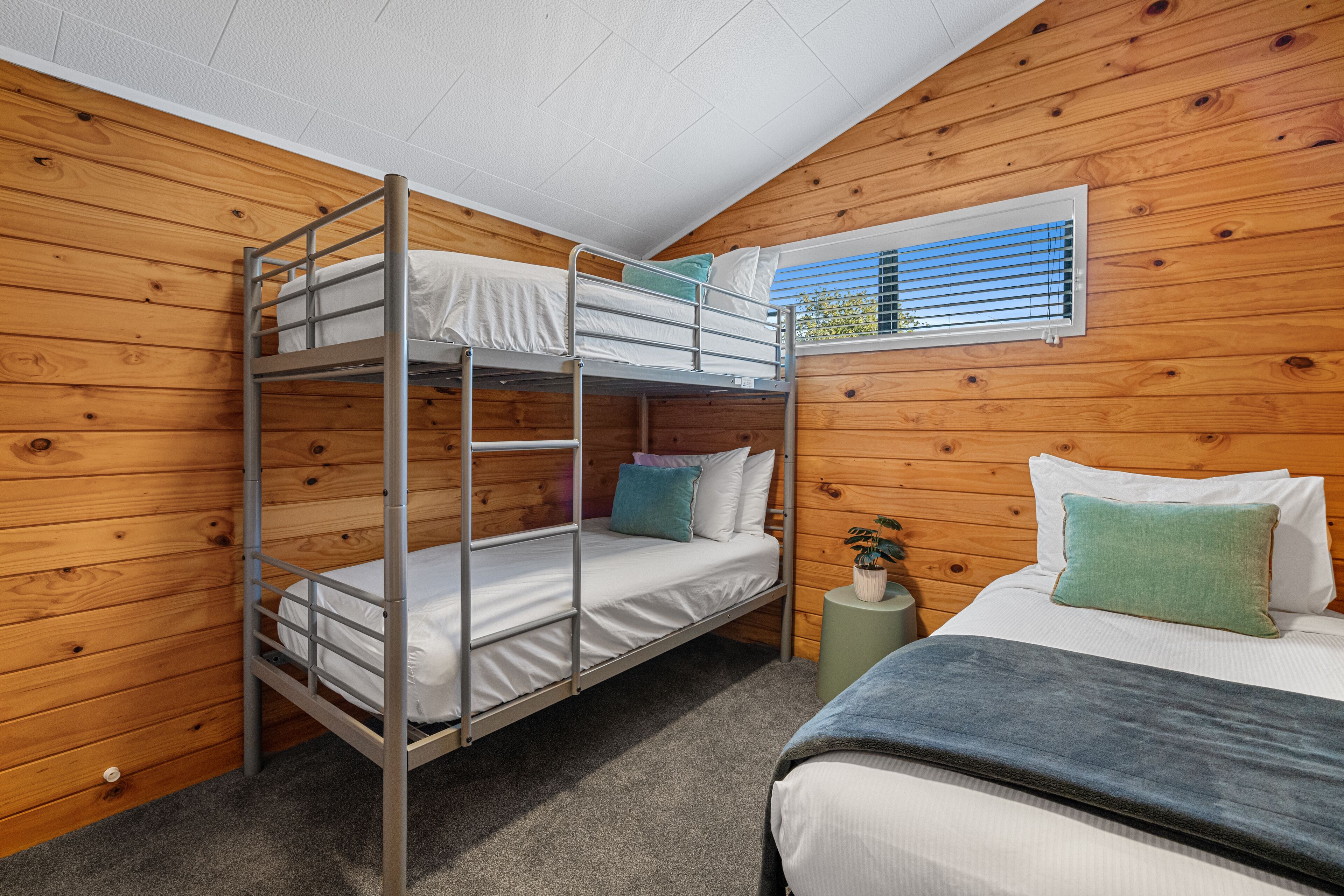 A bunk bed room with wooden walls, a single bed, and a window with blinds.