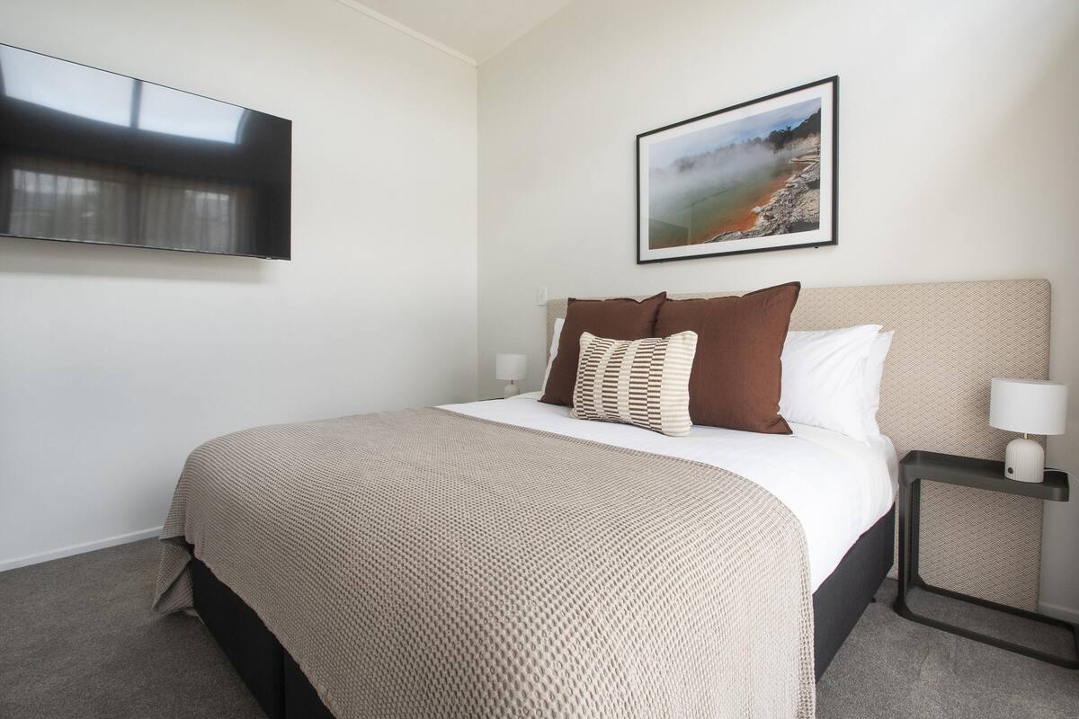Una cama bien hecha con una manta color beige, un almohadón marrón y una almohada blanca. Sobre la cama cuelga un cuadro con una escena de playa. Un televisor de pantalla plana está montado en la pared a la izquierda de la cama.