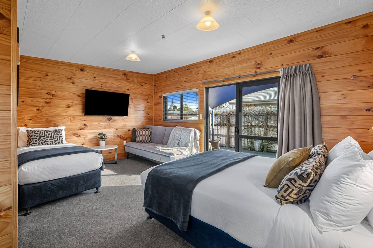 Un dormitorio estilo cabaña moderna con dos camas, un televisor de pantalla plana, un sofá cama y un amplio ventanal corredero al exterior.