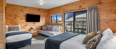 A modern log cabin bedroom with two beds, a flat-screen TV, a sofa bed, and a large sliding glass door to the outside.