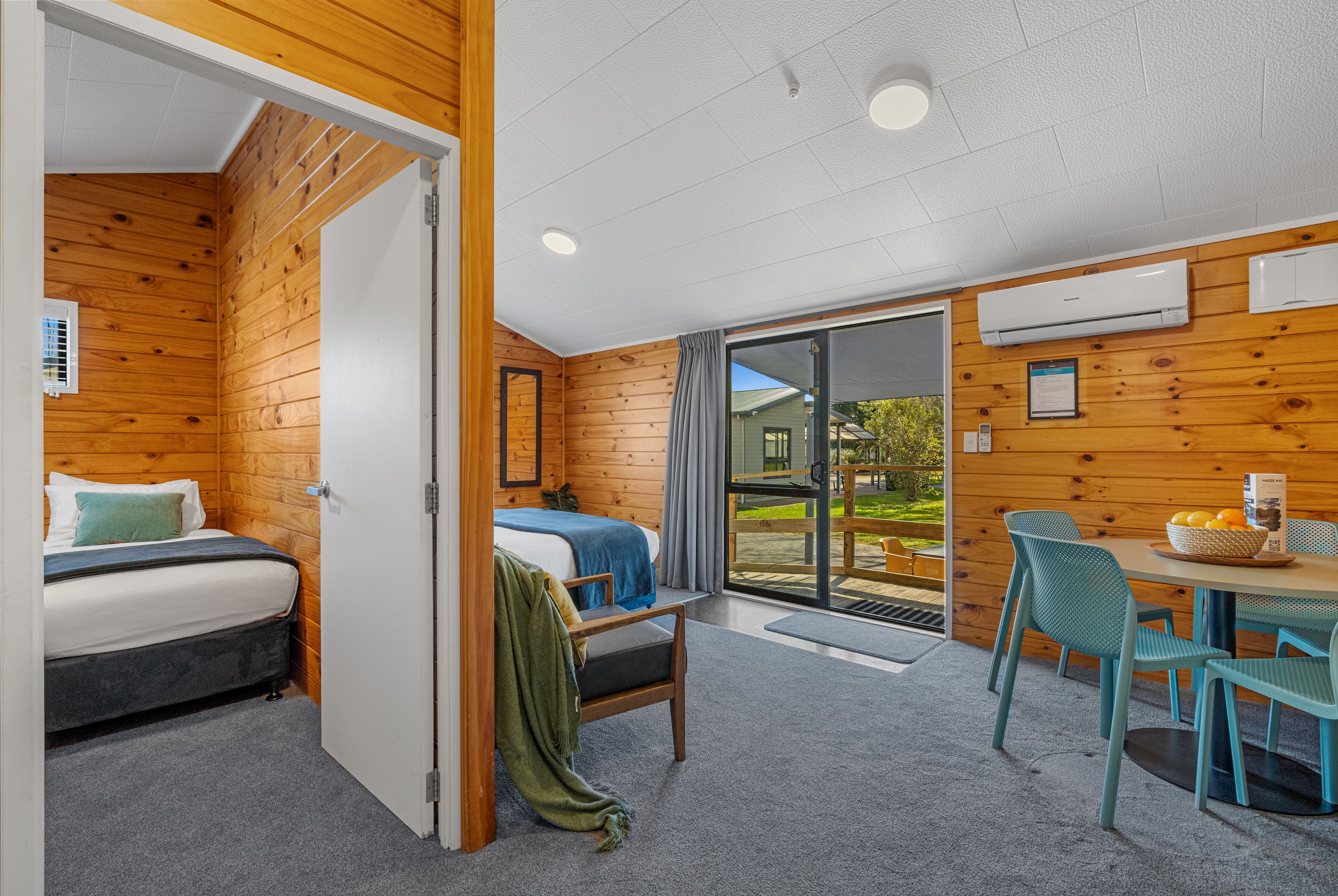 A room with wooden walls, a bed, a desk with chairs, and a view through a glass door.