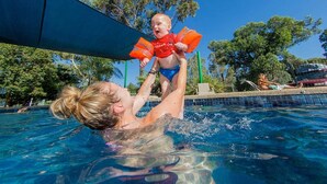 Una piscina con acqua cristallina, una donna e un bambino che giocano, e una persona che si rilassa su un lettino da spiaggia sullo sfondo.