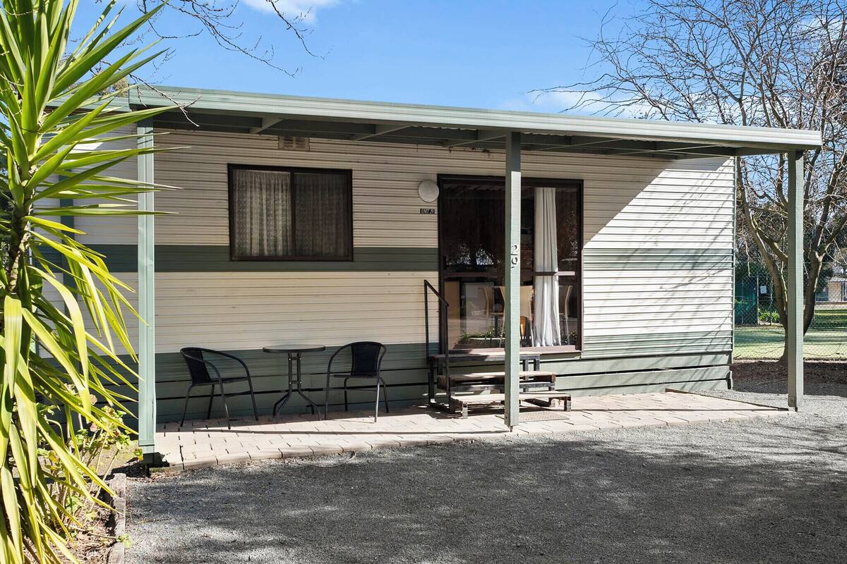 A small, single-story RV with a covered porch, a small table, and two chairs.