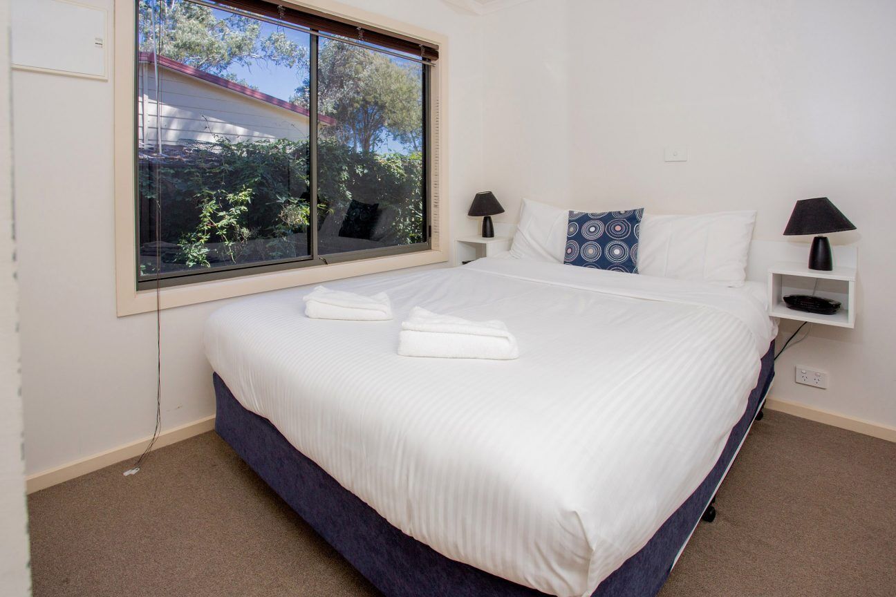 Una cama bien hecha con sábanas blancas y dos almohadas, una ventana con vistas a árboles y una mesita de noche con una lámpara.