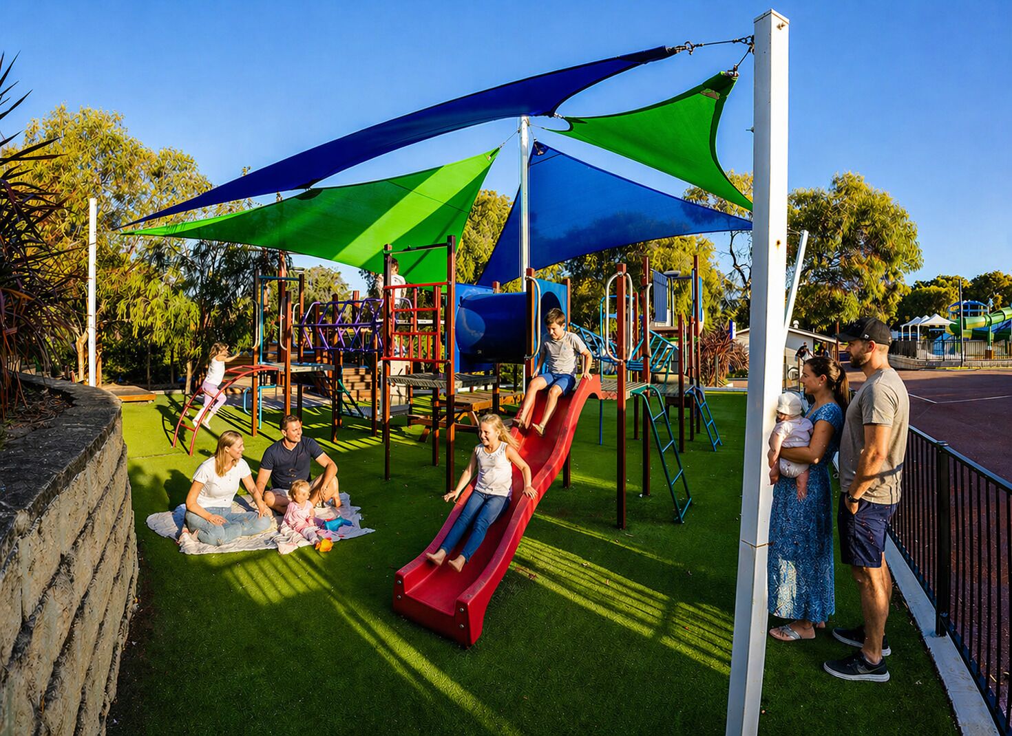 A playground with slides and climbing structures, surrounded by green artificial grass and shaded by large canopies.