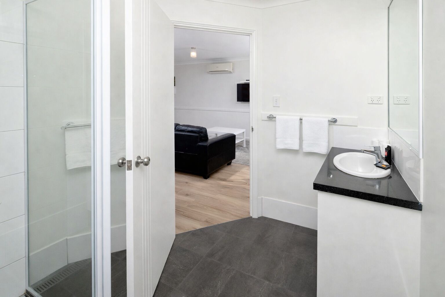 A modern bathroom with a glass shower enclosure, a white sink with a black countertop, and a visible bedroom with a black leather sofa.