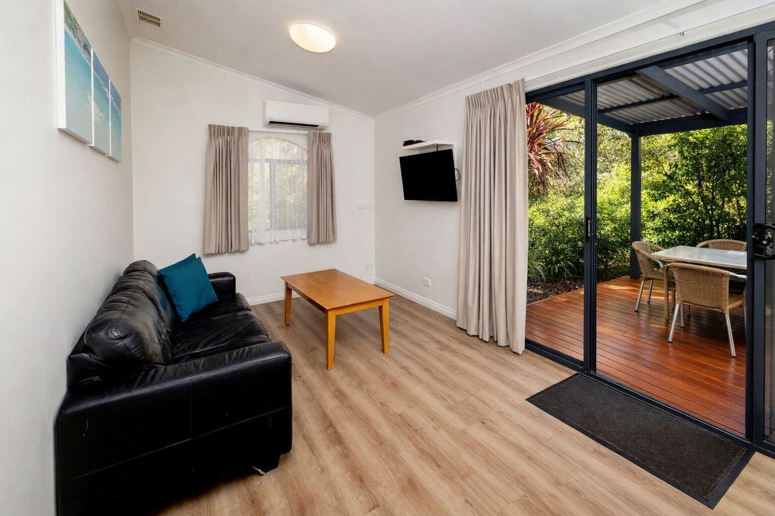 A room with a black leather sofa, a wooden coffee table, a flat-screen TV mounted on the wall, and a sliding glass door leading to an outdoor patio with wicker furniture.