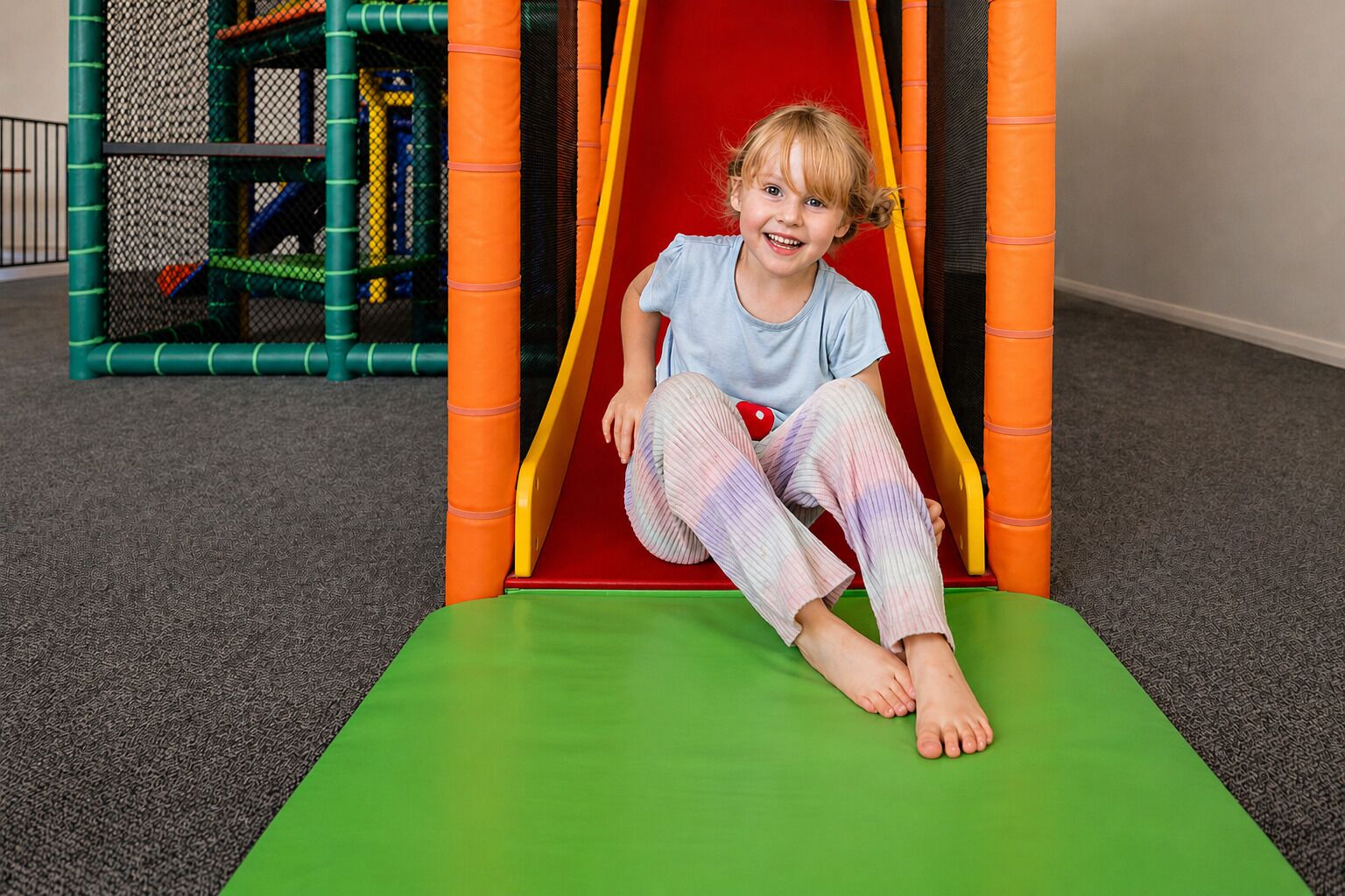 A child sitting on a slide in a play area with a netted structure in the background.