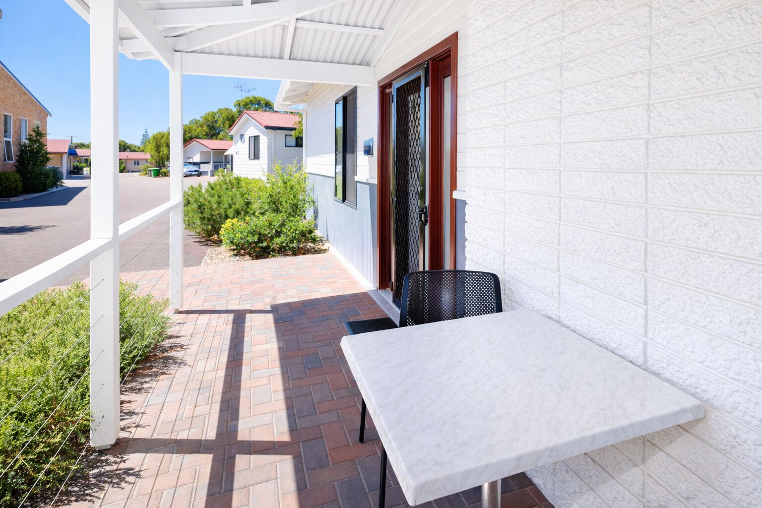 A covered outdoor seating area with a table and chairs, a brick pathway, and a white building with a window and door.