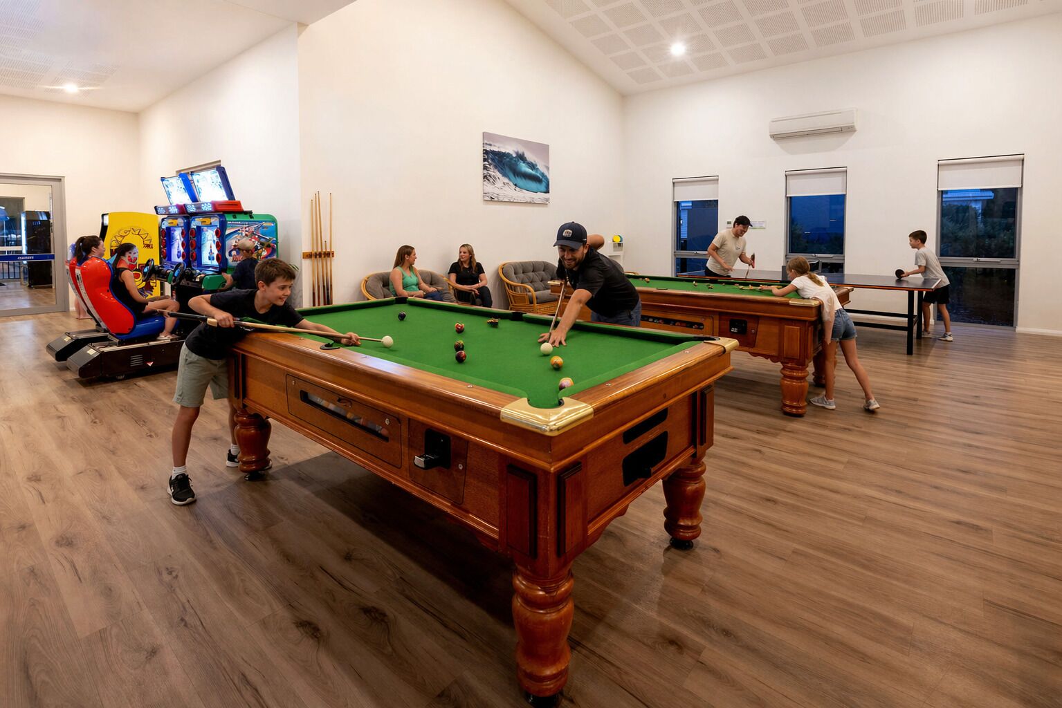 Una sala de billar con múltiples mesas y jugadores, suelos de madera y un interior luminoso.
