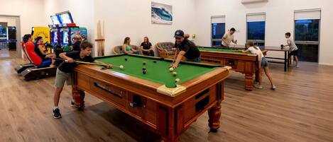 Una sala de billar con múltiples mesas y jugadores, suelos de madera y un interior luminoso.