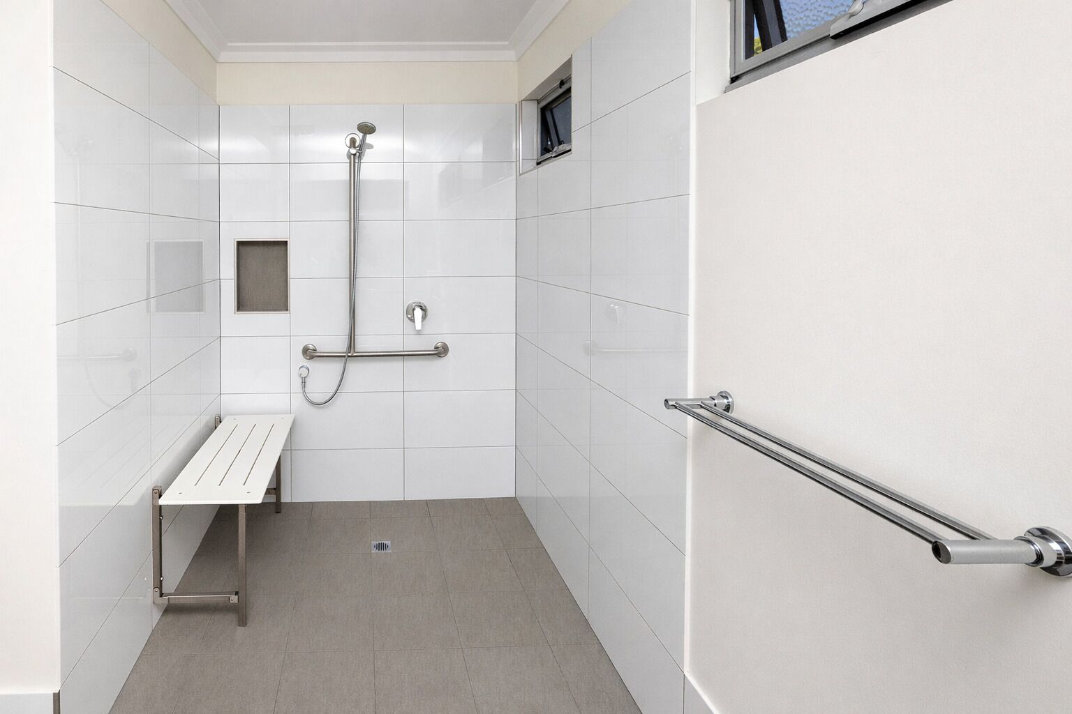 A bathroom with white tiled walls, a bench, a handrail, and a shower with a handheld showerhead.