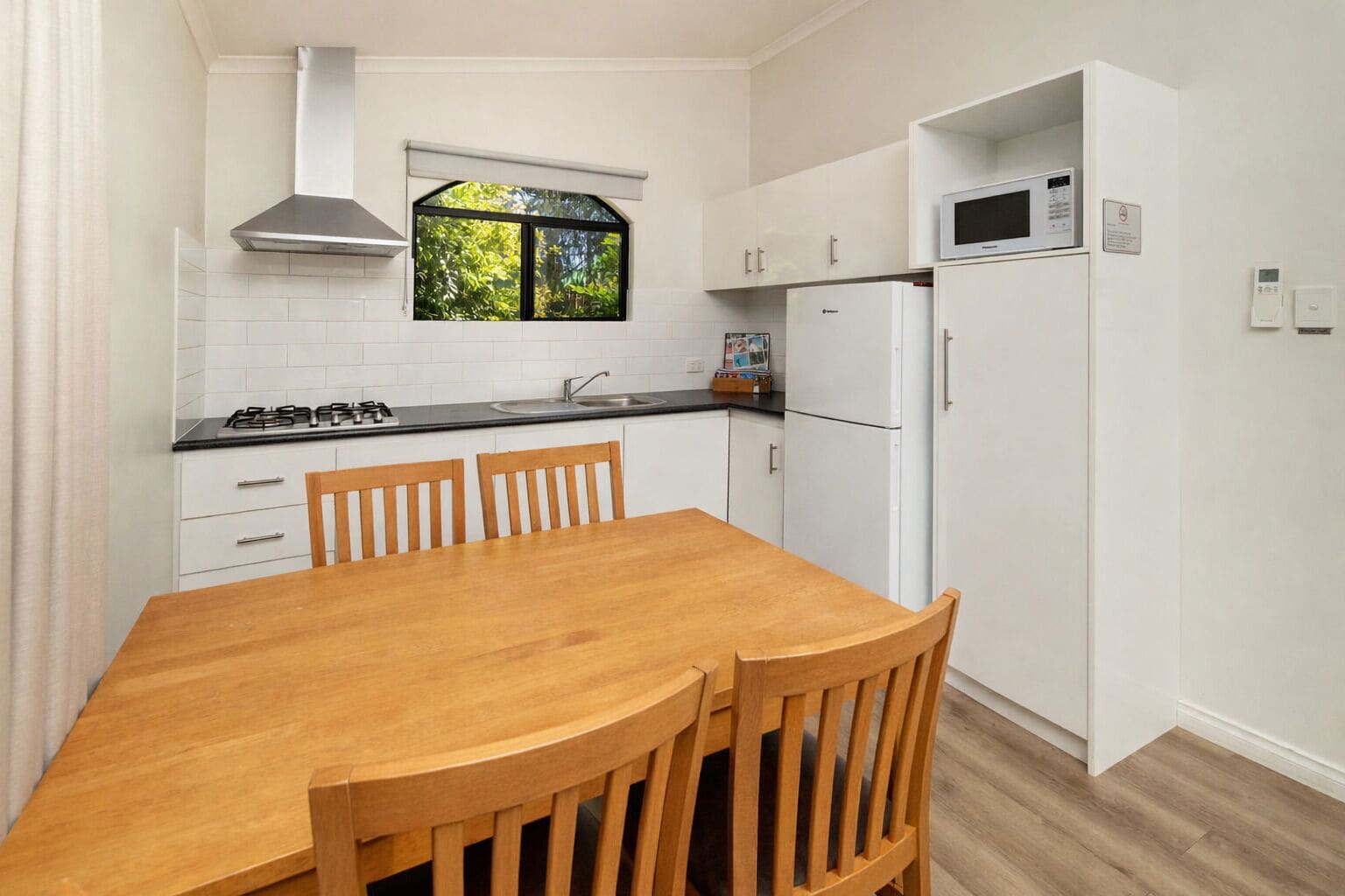 Cocina compacta con armarios blancos, una mesa de comedor de madera y una ventana con vistas a la vegetación.