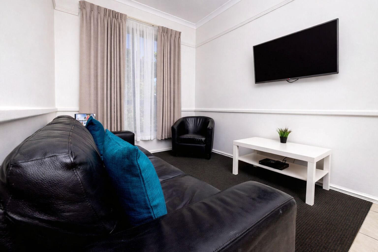 A modern living room with a black leather sofa, a white side table, a black leather chair, and a mounted TV.