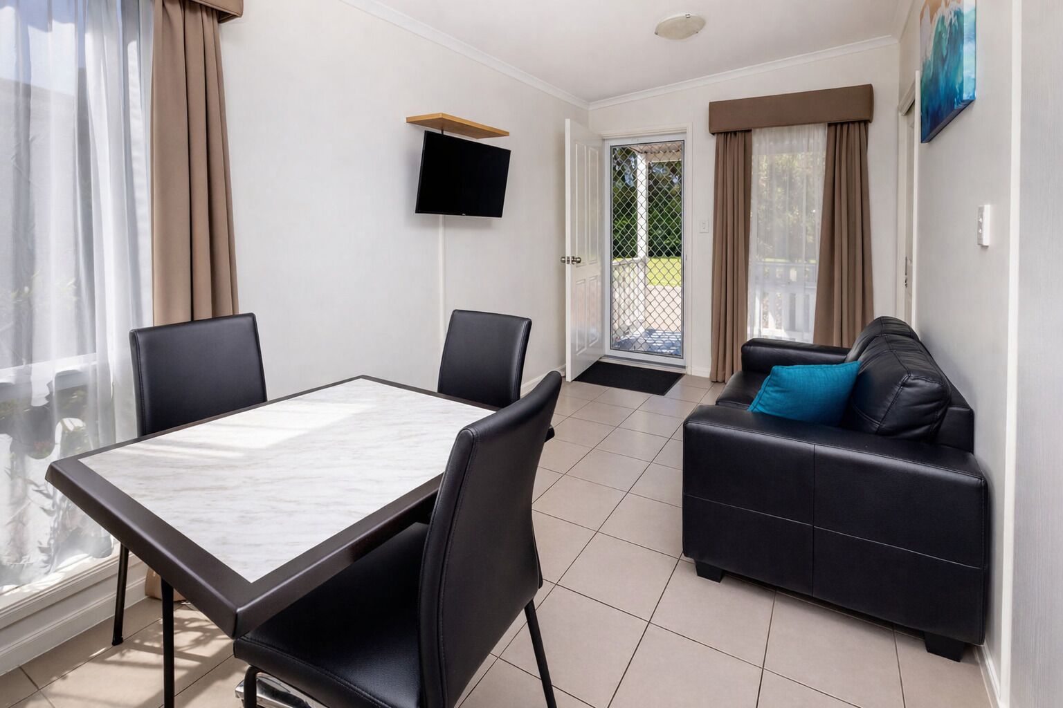 Een moderne kamer met een eettafel, stoelen, een zwarte leren bank, een aan de muur bevestigde televisie en een schuifdeur van glas naar een buitenruimte.