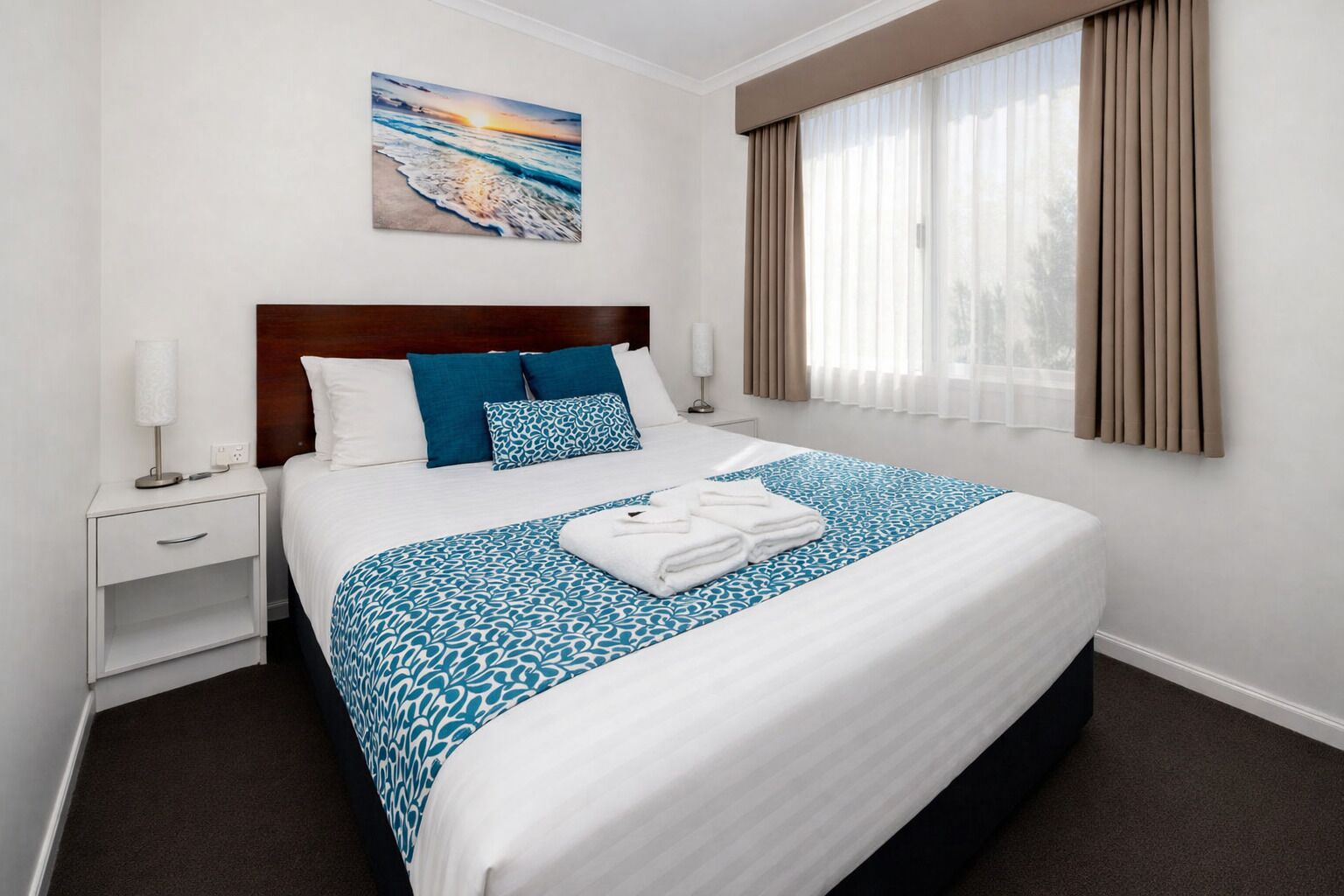 A hotel room with a large bed, a nightstand, a lamp, a painting of a beach scene, and a window with curtains.