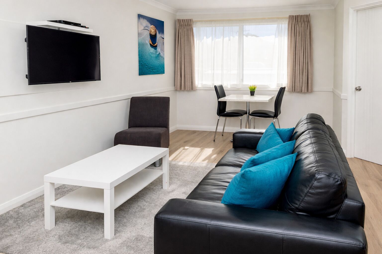 A living room with a black leather sofa, a white coffee table, a grey armchair, a flat-screen TV mounted on the wall, a dining table with chairs, and a window with curtains.