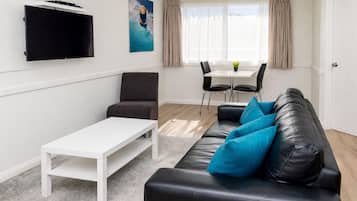 A living room with a black leather sofa, a white coffee table, a grey armchair, a flat-screen TV mounted on the wall, a dining table with chairs, and a window with curtains.
