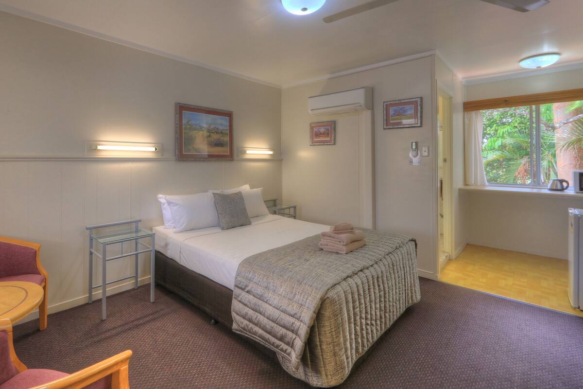 Habitación de hotel con cama, una silla, una mesita y ventana con vistas a zonas verdes.