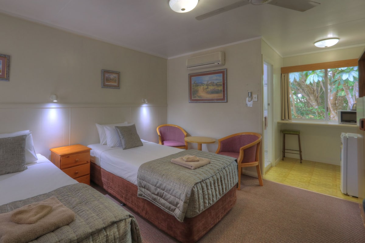 Habitación de hotel con dos camas, un escritorio, una silla y una ventana con vistas a árboles.